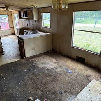 Gutted mobile-home interior after tear-out: flooring stripped to subfloor, cabinets and countertop removed, bright windows