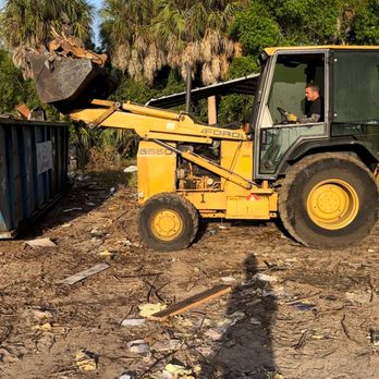 The client's own yellow Ford backhoe loader with the operator in the cab, bucket raised over a green roll-off dumpster