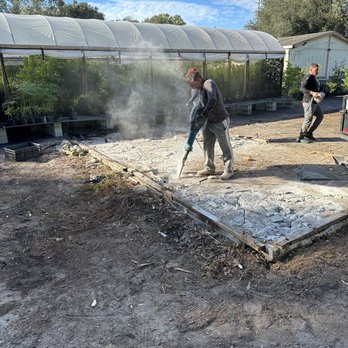 Two workers breaking a concrete slab with a jackhammer on a nursery job site, dust plume rising, greenhouse structure behind