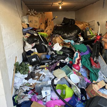 Interior of a packed storage unit, floor-to-ceiling mound of bags, clothing and housewares under a bare ceiling light