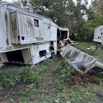 Two derelict, gutted travel trailers rusting in overgrown brush with a torn sheet-metal panel on the ground